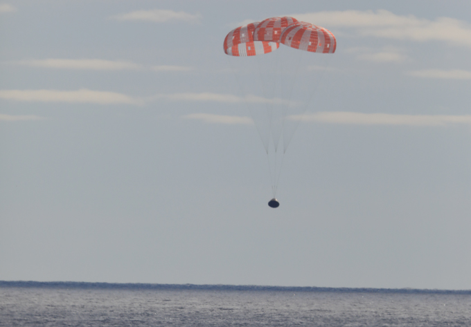 la nave espacial Orión desciende hacia el Océano Pacífico, frenada por un gran paracaídas rojo y blanco